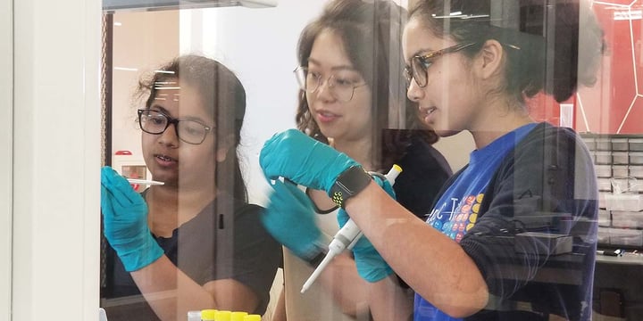 three girls wearing blue gloves do something on the other side of a window