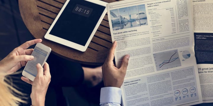 hands of two people at a table, one reading a news letter, the other looking at a smartphone, a tablet unused on the table