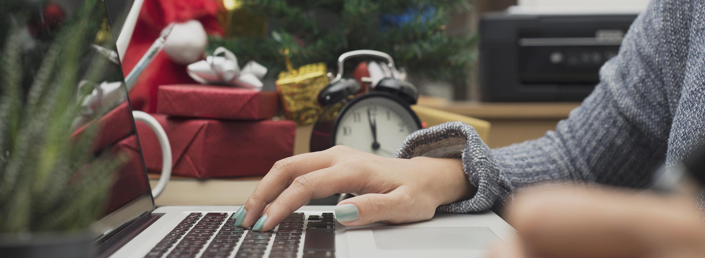 hand on laptop computer; in background, wrapped gifts and a clock with hands nearly at 12