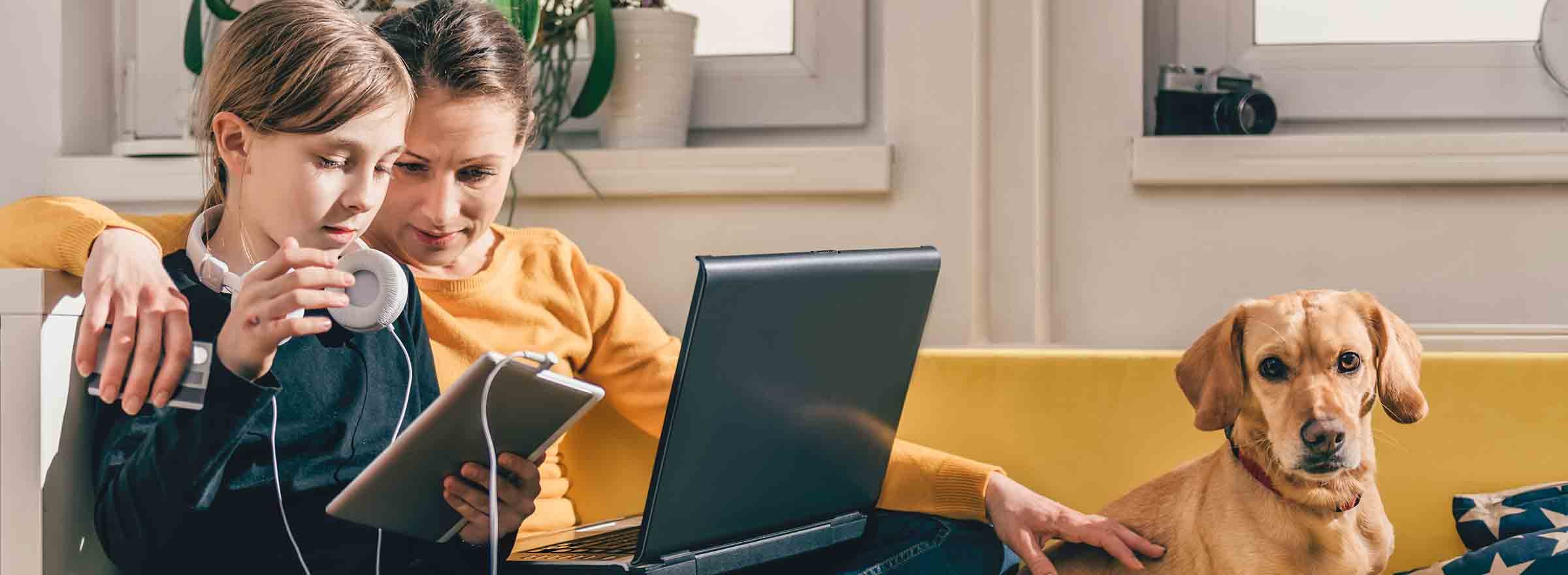 parent, child, and dog on a sofa; a laptop is open, and the child is looking at a tablet but not listening to the attached headphones; the parent holds a credit card