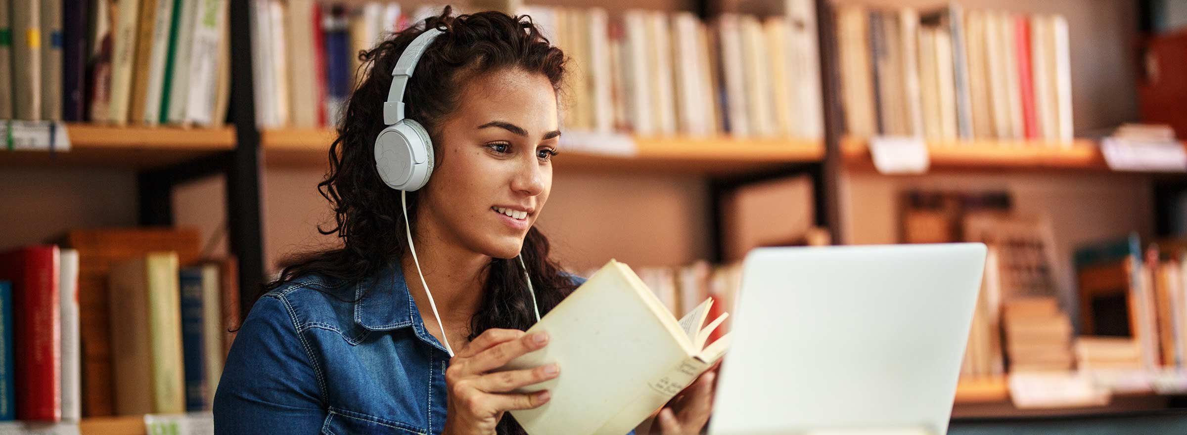 woman in library with laptop, headphones, and a paper book