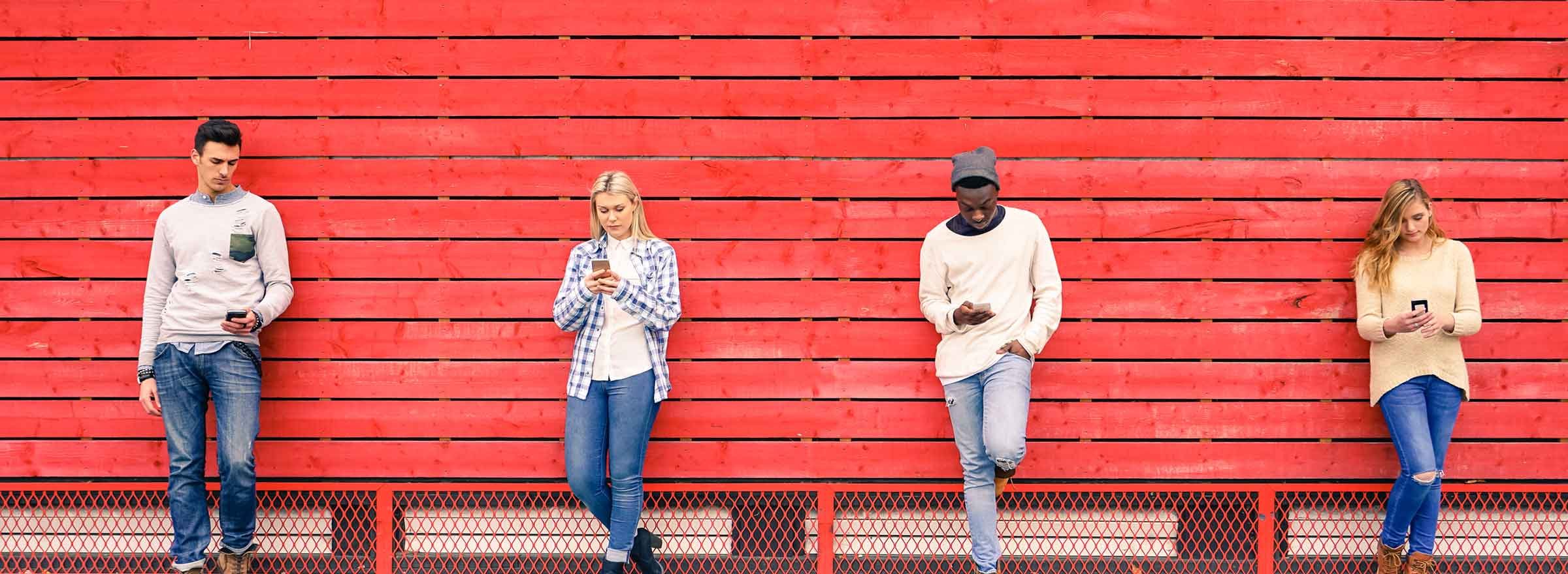 four people leaning against a red fence and looking at smartphones