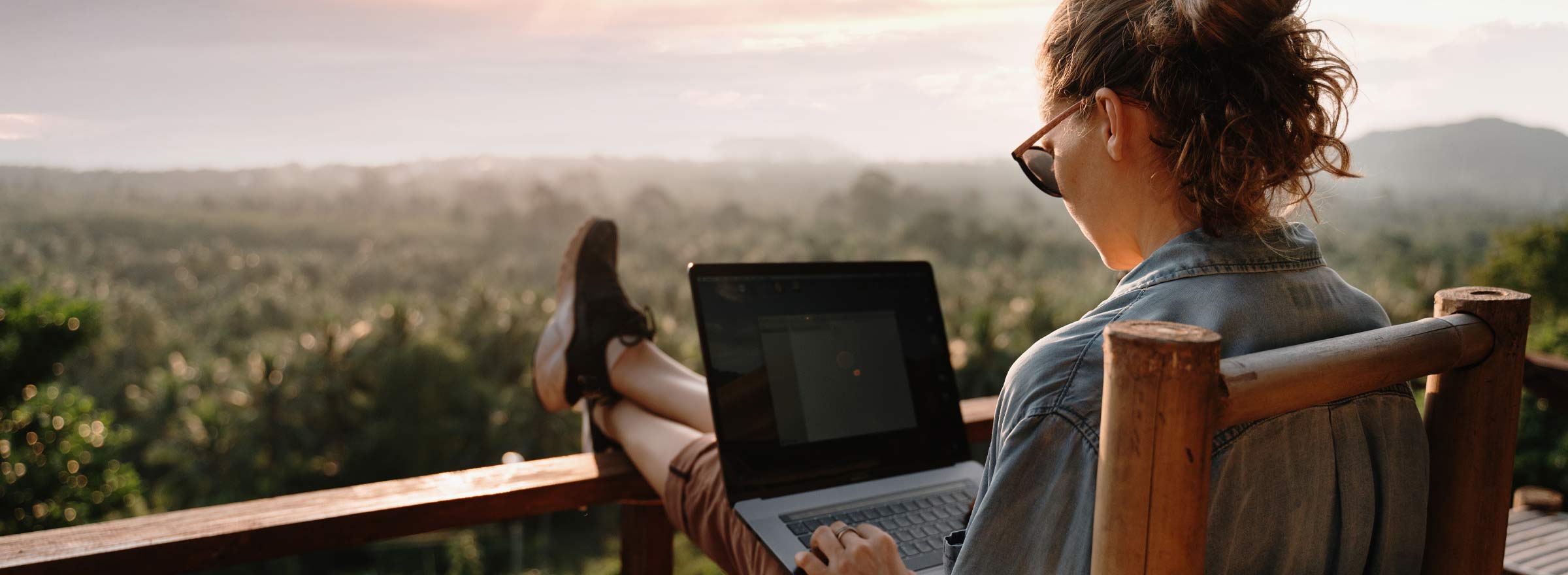 woman using a laptop, feet up on a railing, overlooking a treescape