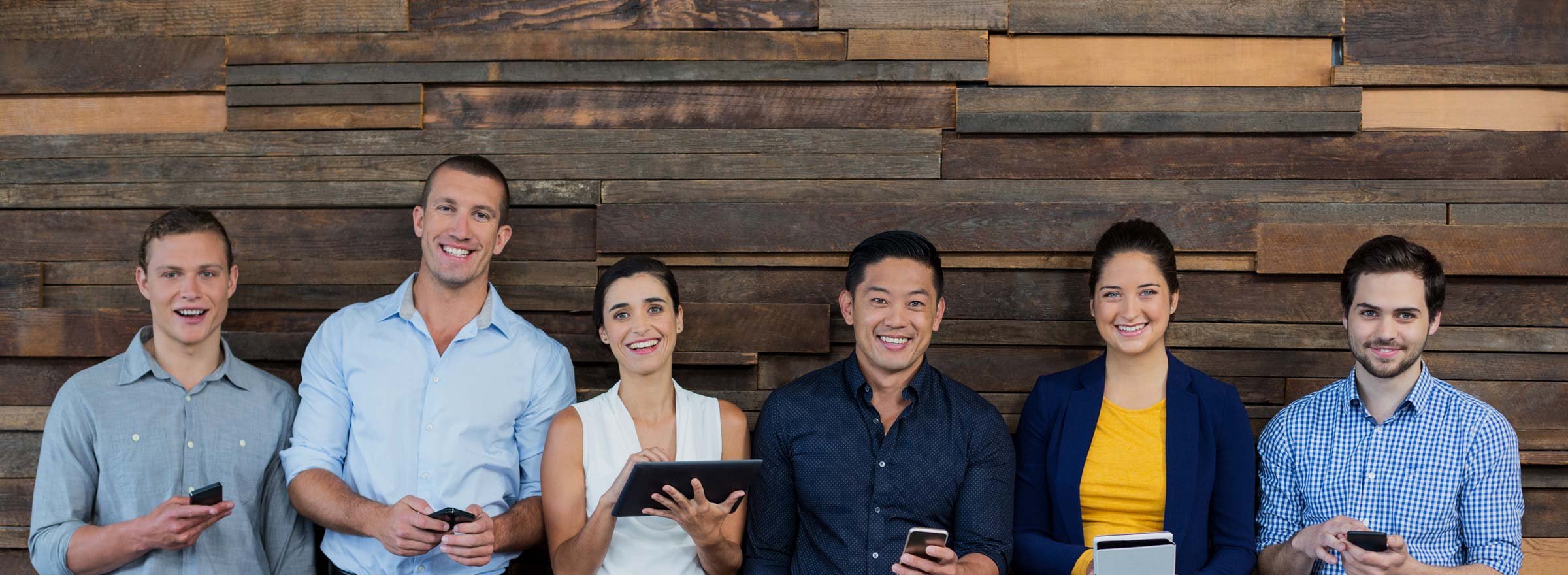 two women and four men standing against a wooden wall, holding cellphones or tablets, and smiling