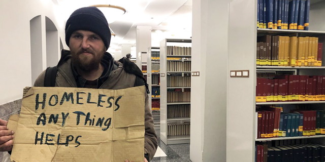 Man holding a sign that reads 'Homeless Anything Helps' inside of a library
